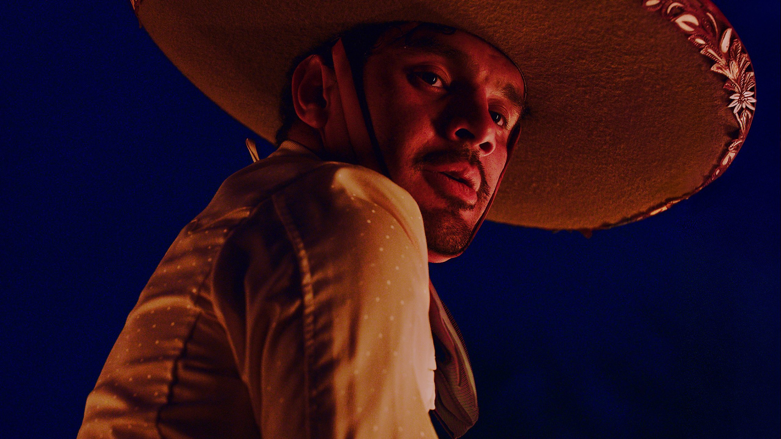 A man wearing a large, decorated sombrero and a tan, dotted shirt looks over his shoulder against a dark blue background.