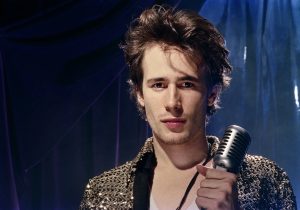 A young man with tousled hair holds a vintage microphone, wearing a sequined jacket, standing under stage lights with a dark curtain in the background.