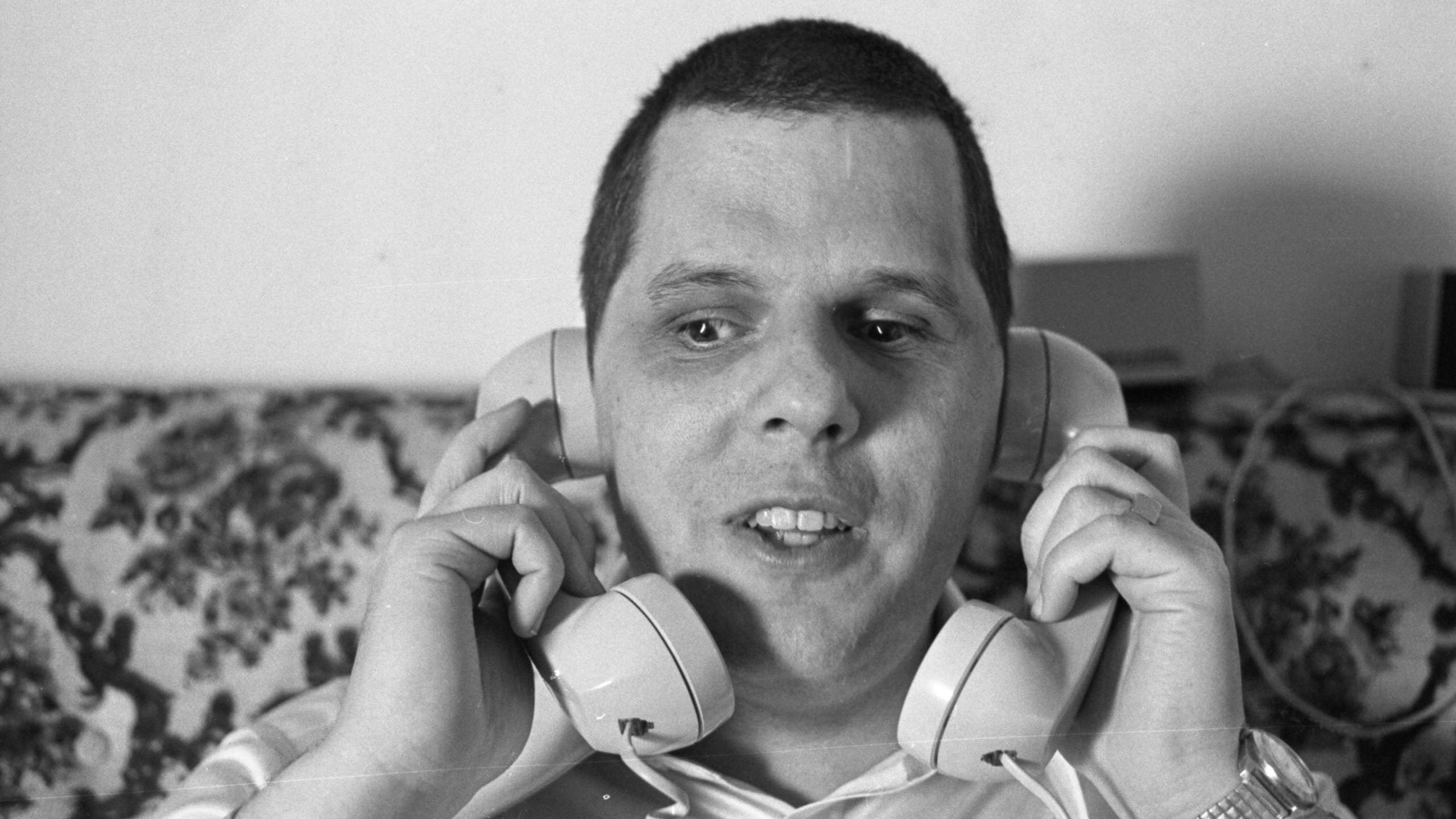 A man sits on a patterned couch, holding two telephone receivers to his ears, looking slightly to the side.