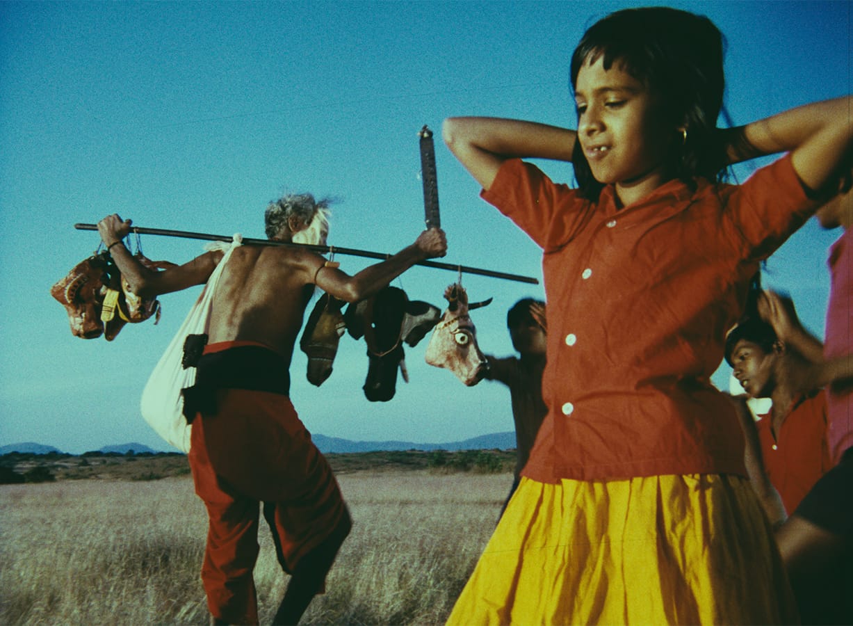 A group of children, including a girl in a yellow skirt, stands in a field while an older man carries a stick with objects hanging from it.