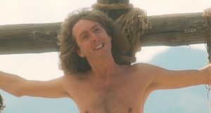 A shirtless person with long hair smiles while tied to a wooden cross outdoors, with blue sky and mountains in the background.
