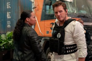 A woman and a man in tactical gear stand together outdoors near a truck, both looking alert and serious.