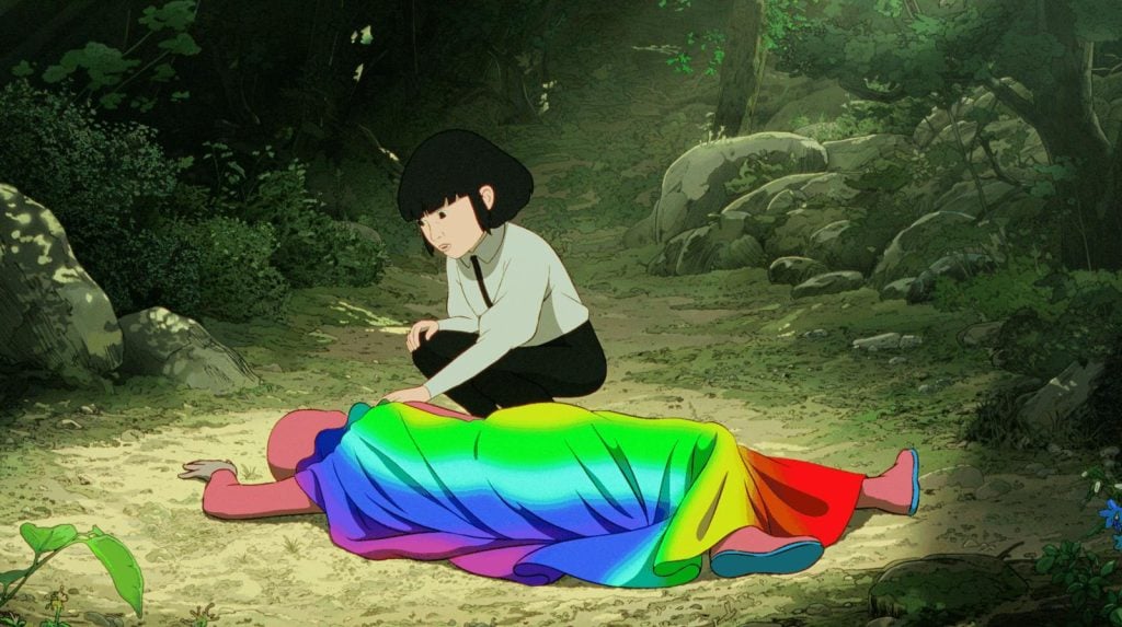 A person with short black hair kneels beside another figure lying on the ground, covered with a rainbow-colored blanket, in a forest setting.