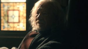 An elderly man with long white hair and a beard sits in a dimly lit room beside a stained glass window, wearing a dark robe.