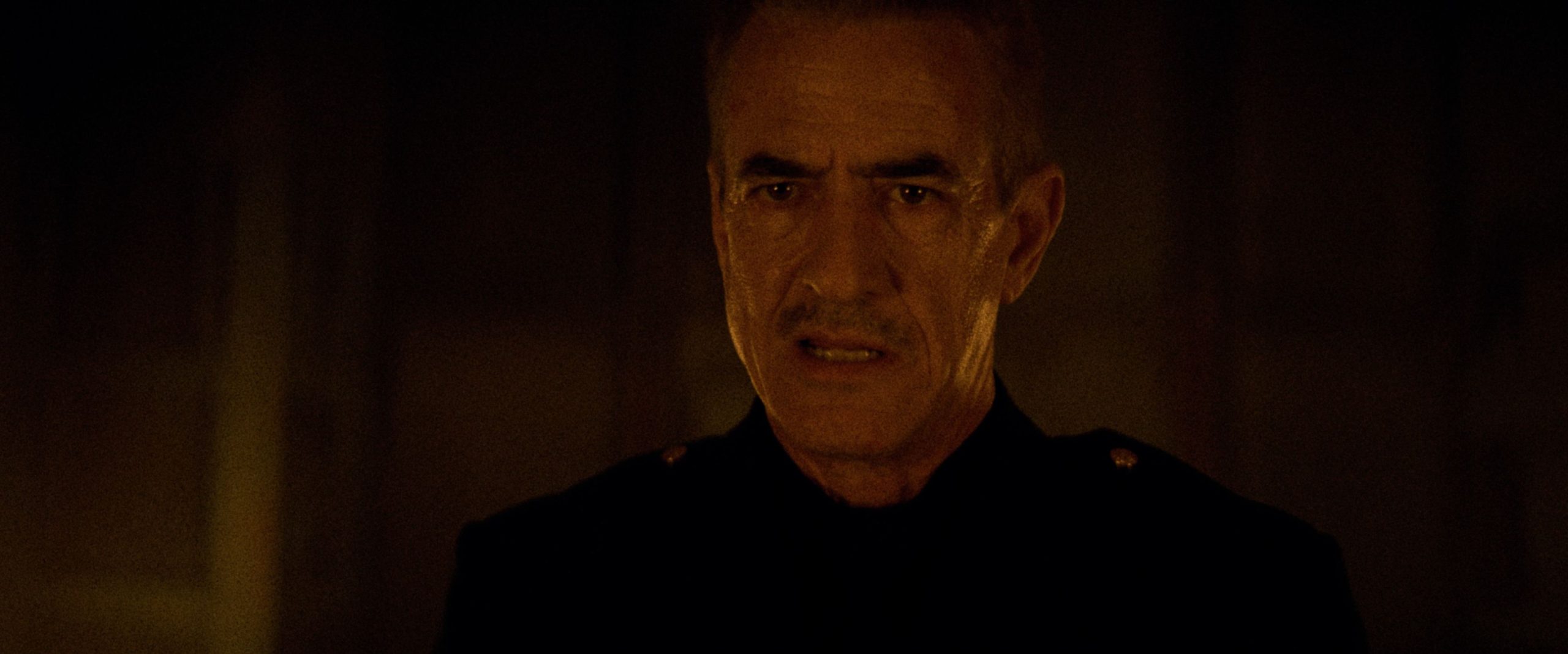 A middle-aged man with short gray hair and a mustache, wearing a dark uniform, looks ahead with a serious expression in dim lighting.