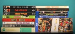 A stack of DVD and Blu-ray cases, including titles like Under Siege, Evil Dead Rise, Snakes on a Plane, Shameless, and Two and a Half Men, arranged on a teal surface.