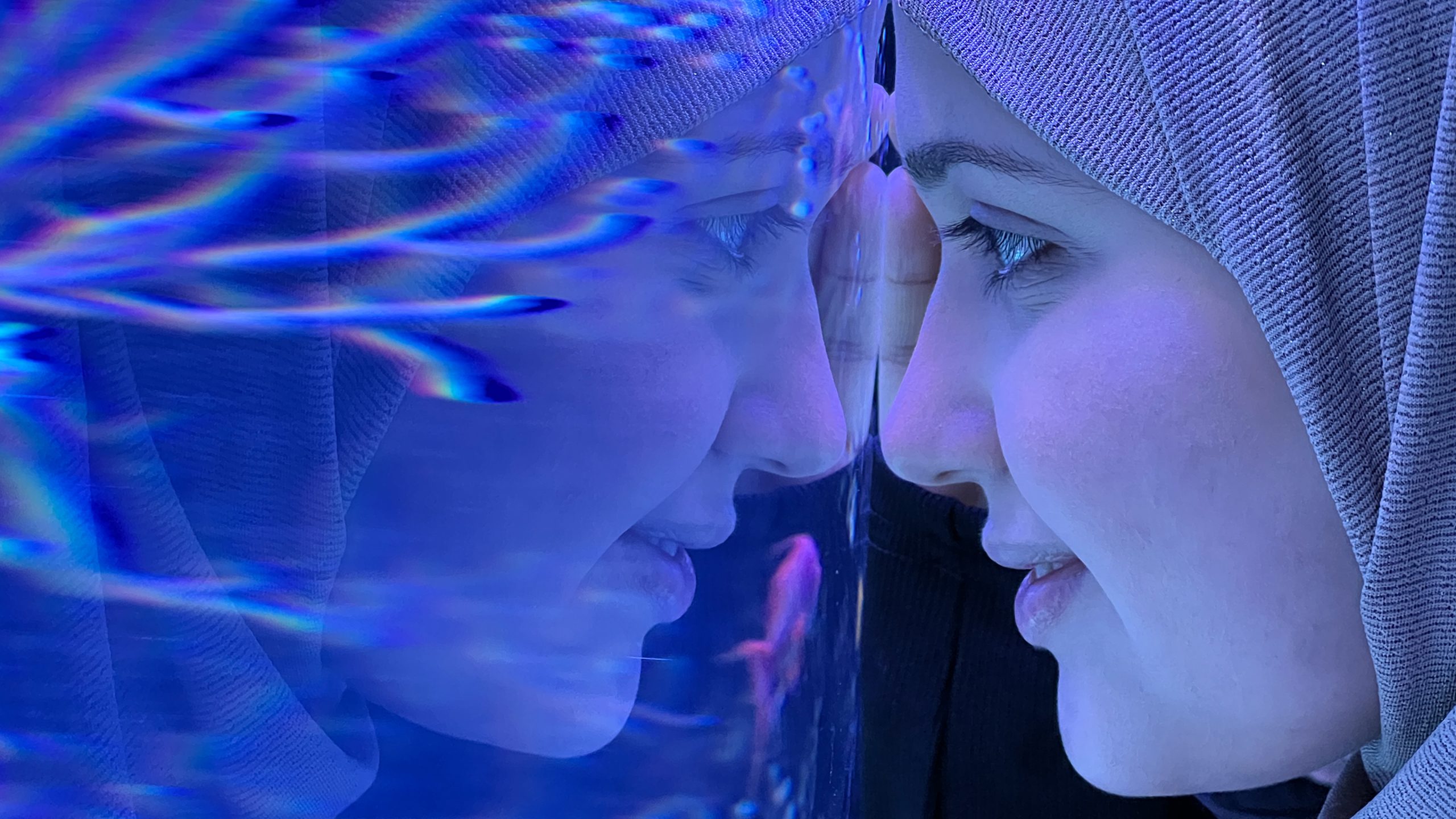 A person in a headscarf smiles closely at an aquarium glass, their face reflected clearly, with blue water and fish visible in the background.