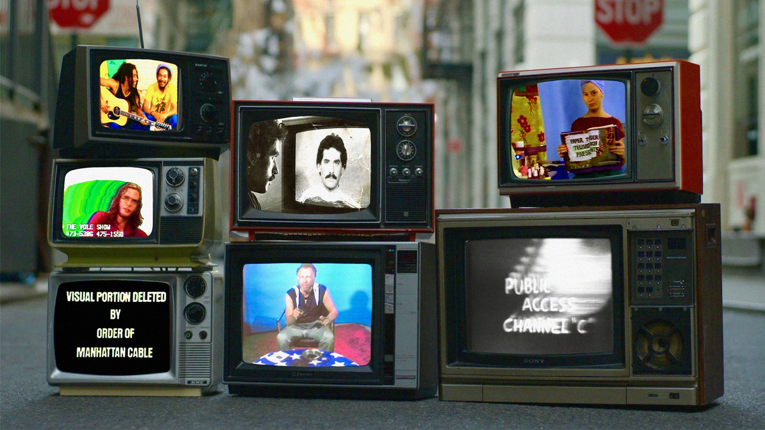 A group of old television sets display various public access TV programs and static, stacked together on a city street.