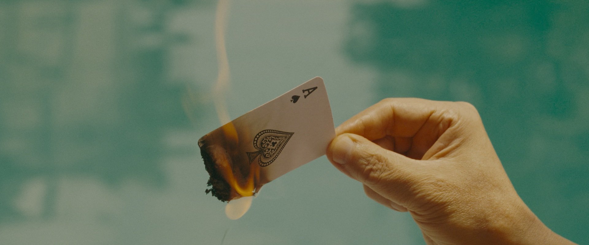 A hand holds an ace of spades playing card that is burning at one corner, with smoke rising against a blurred blue-green background.