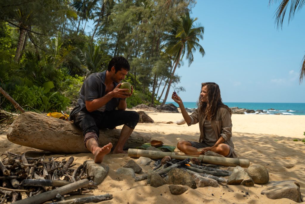 A man and a woman sit on a sandy beach near a campfire, surrounded by tropical trees, as the man drinks from a coconut and the woman gestures while talking.