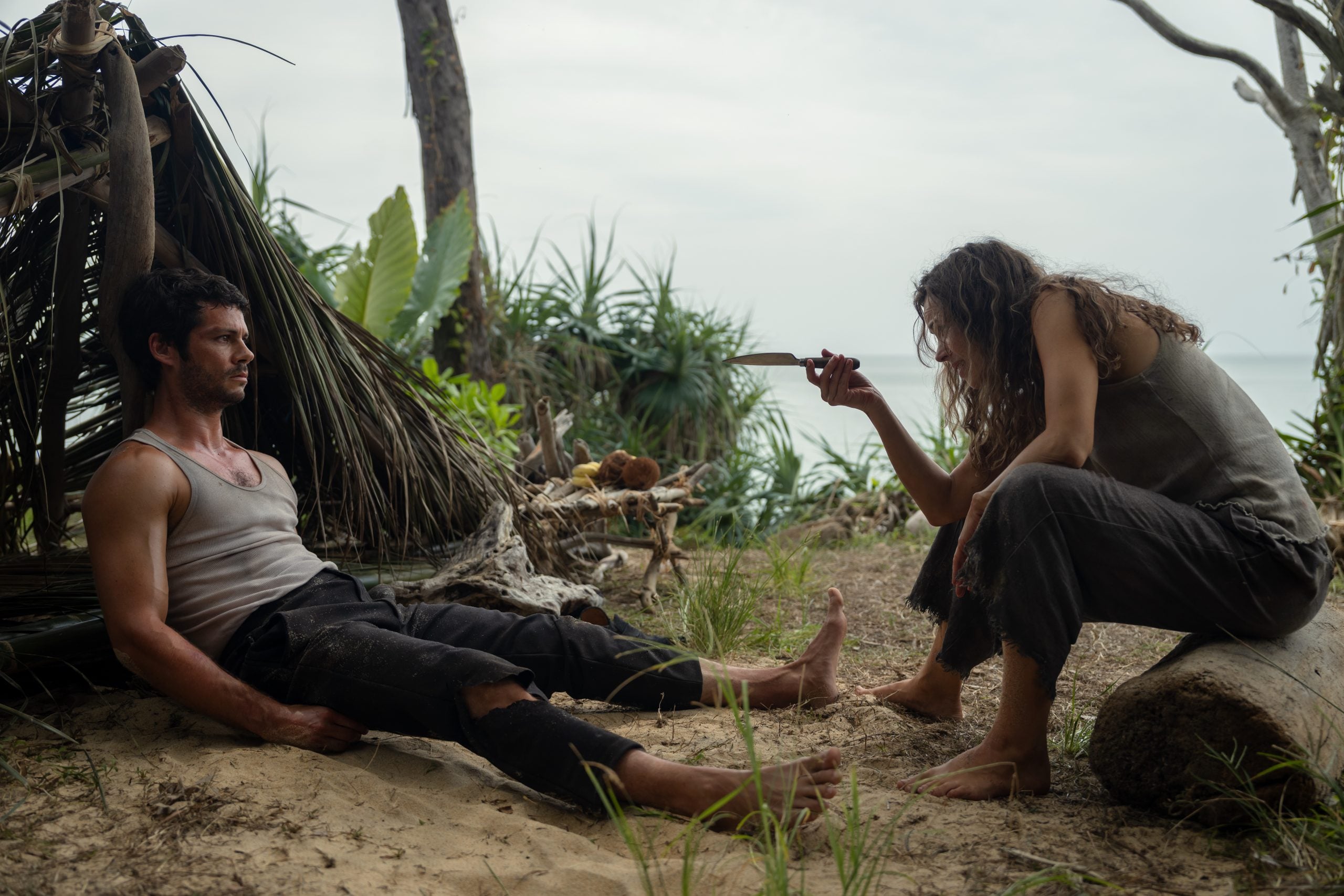 A woman crouches on a log pointing a knife at a man sitting on the ground near a makeshift shelter in a tropical, forested area.
