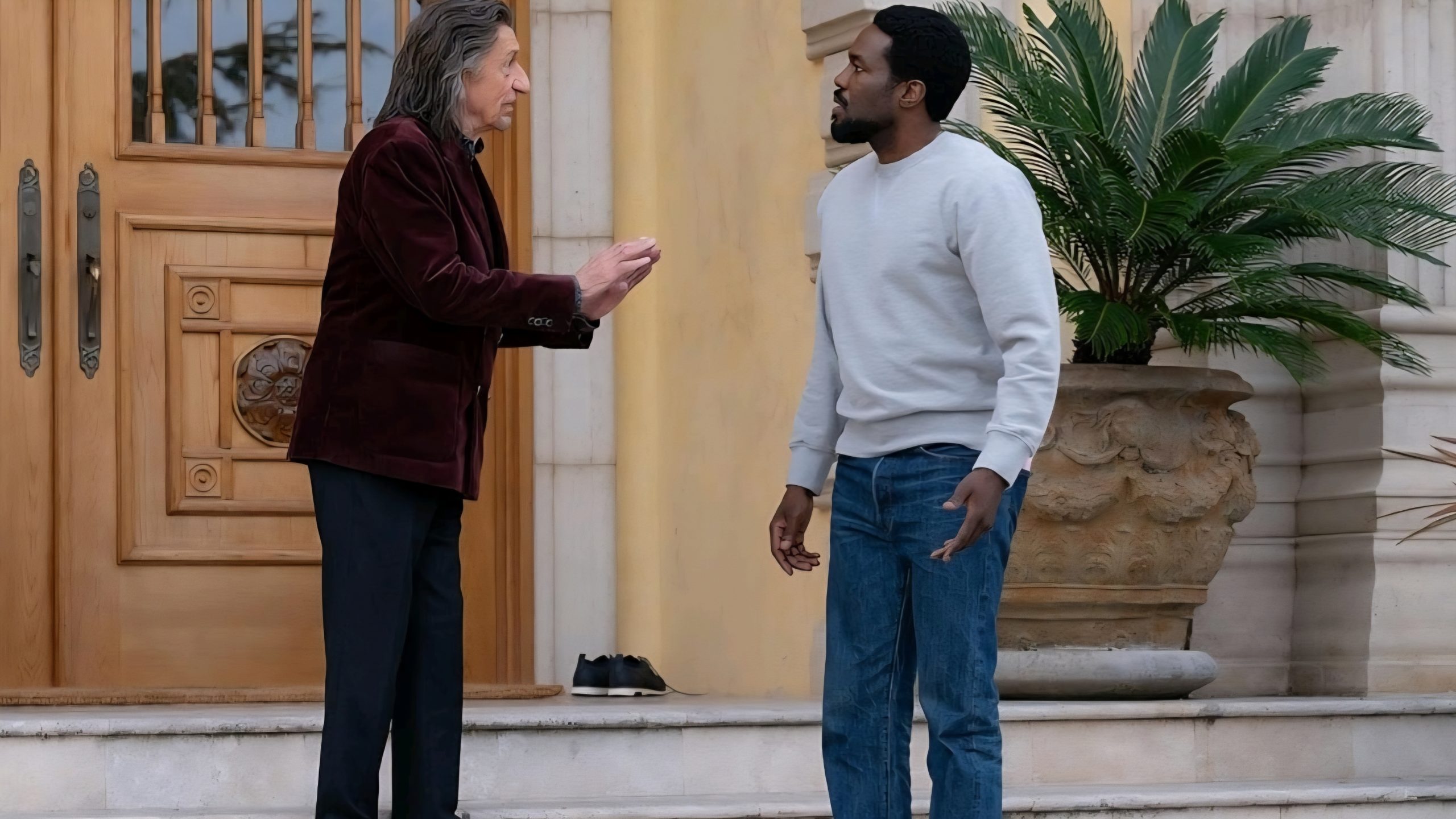 Two men stand and talk on the steps outside a building with large doors and a potted plant; one man gestures with his hands, and a pair of shoes sits on the step behind them.