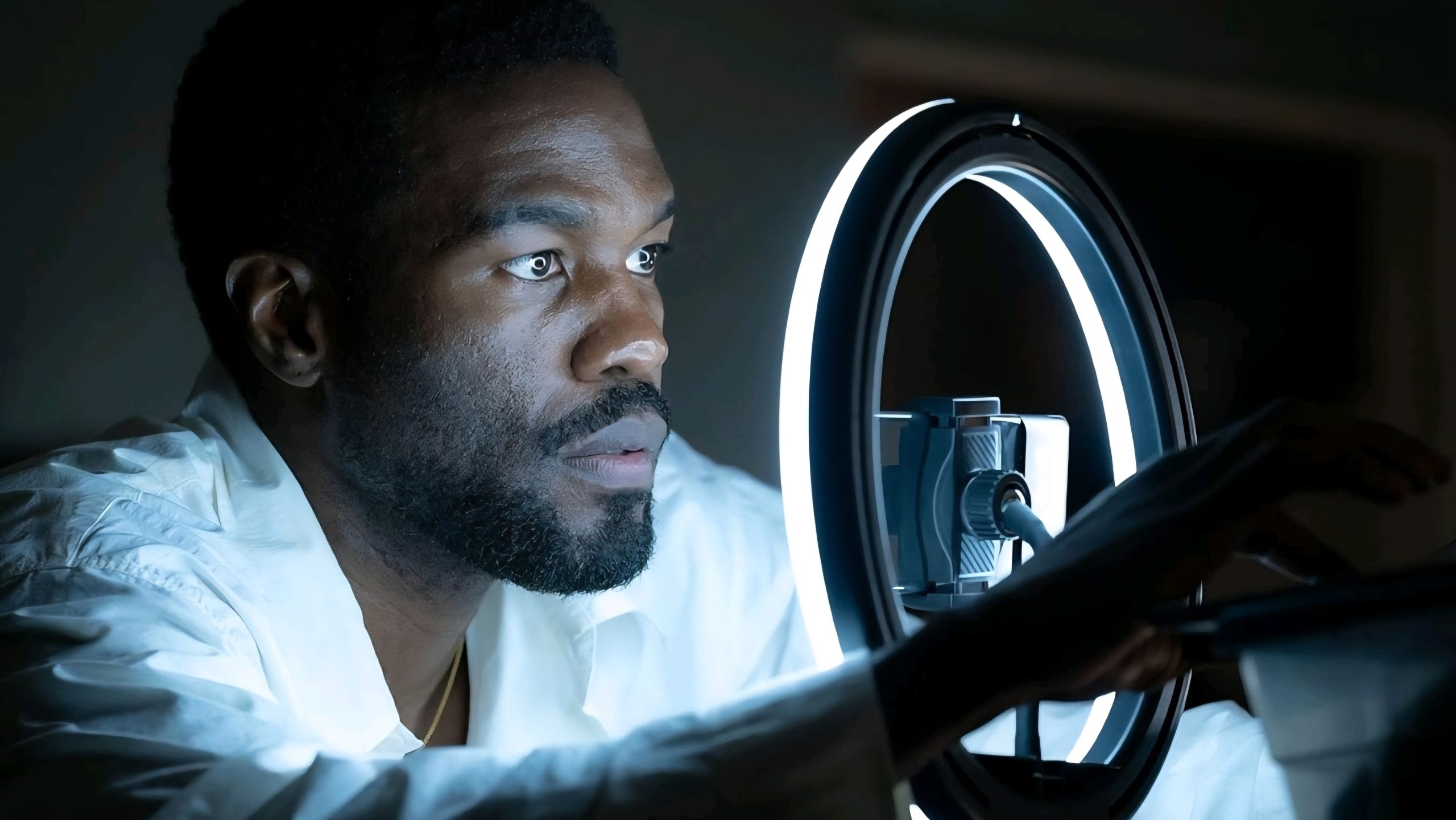 A man in a white shirt adjusts a smartphone positioned inside a lit ring light, focusing on the device in a dimly lit room.