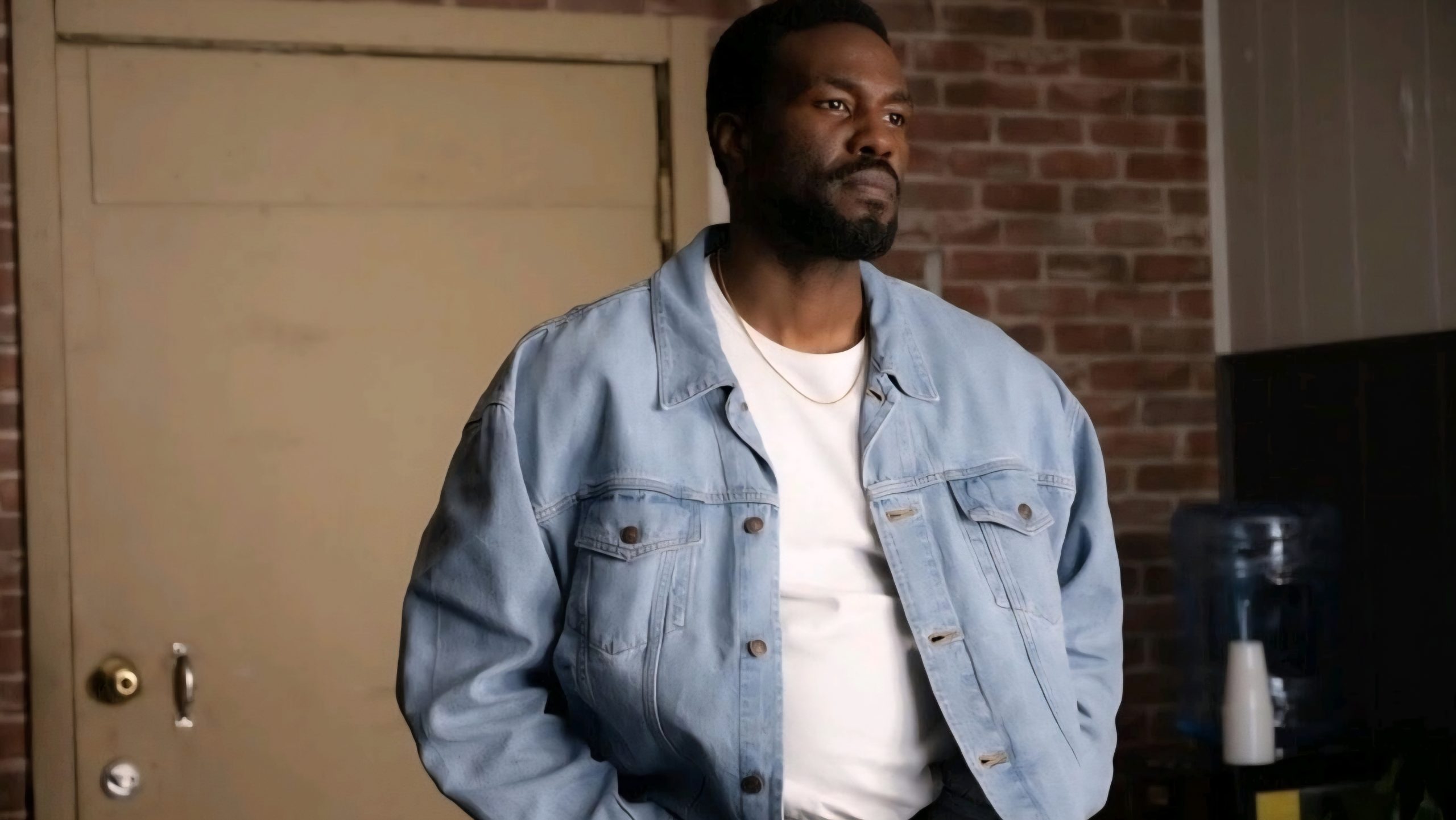 A man wearing a light blue denim jacket and white shirt stands indoors in front of a closed door and brick wall, looking to the side.