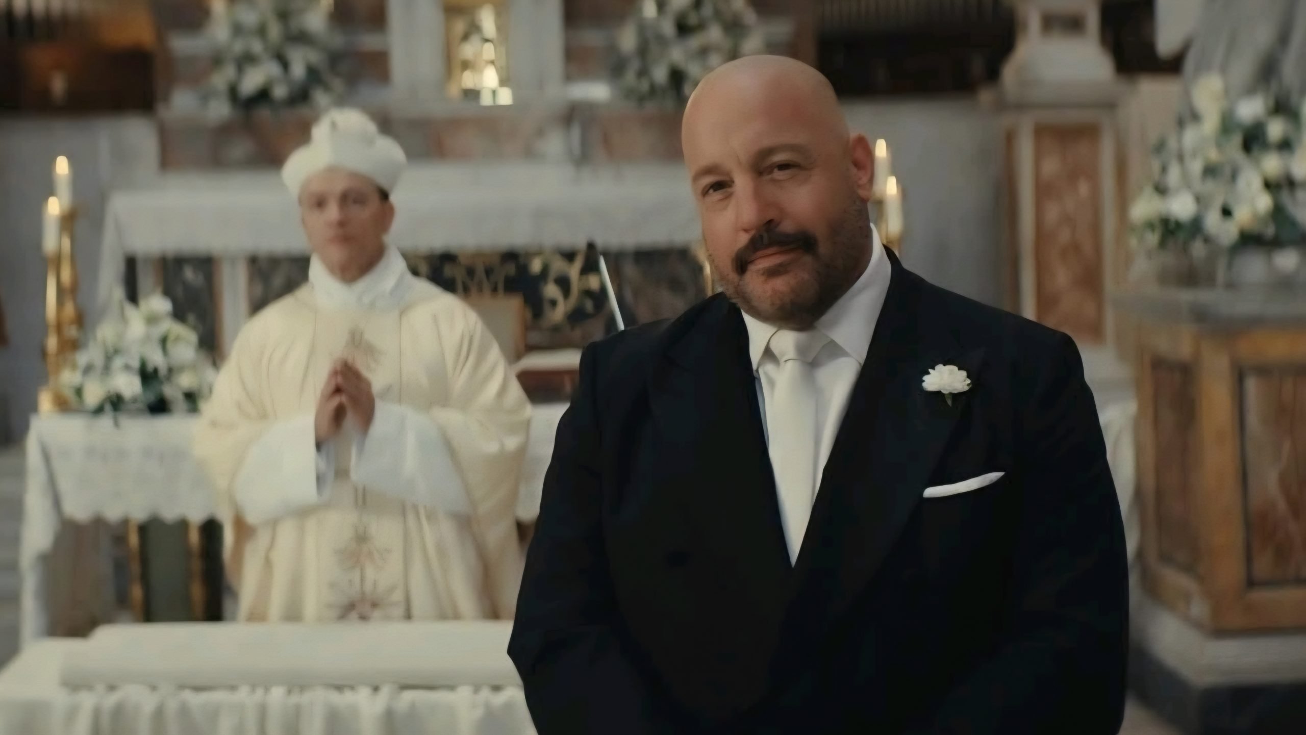 A man in a suit stands in the foreground of a church, with a priest in white robes and floral decorations visible in the background.