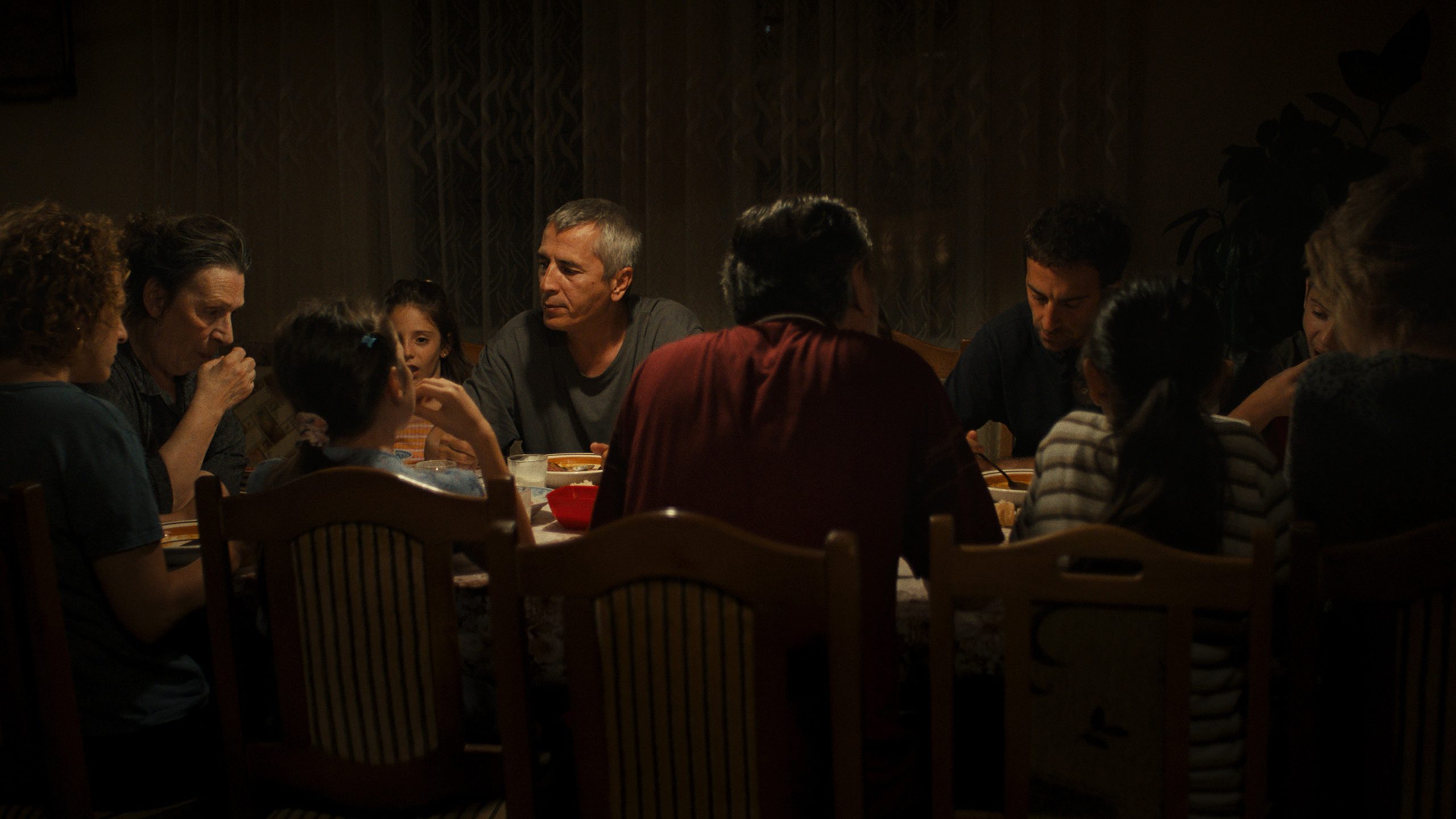 A group of people sits around a dining table, sharing a meal in a dimly lit room.