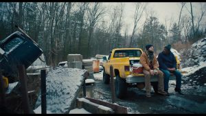 Two men sit on the tailgate of a yellow pickup truck in a snowy, wooded area surrounded by various items and construction materials.