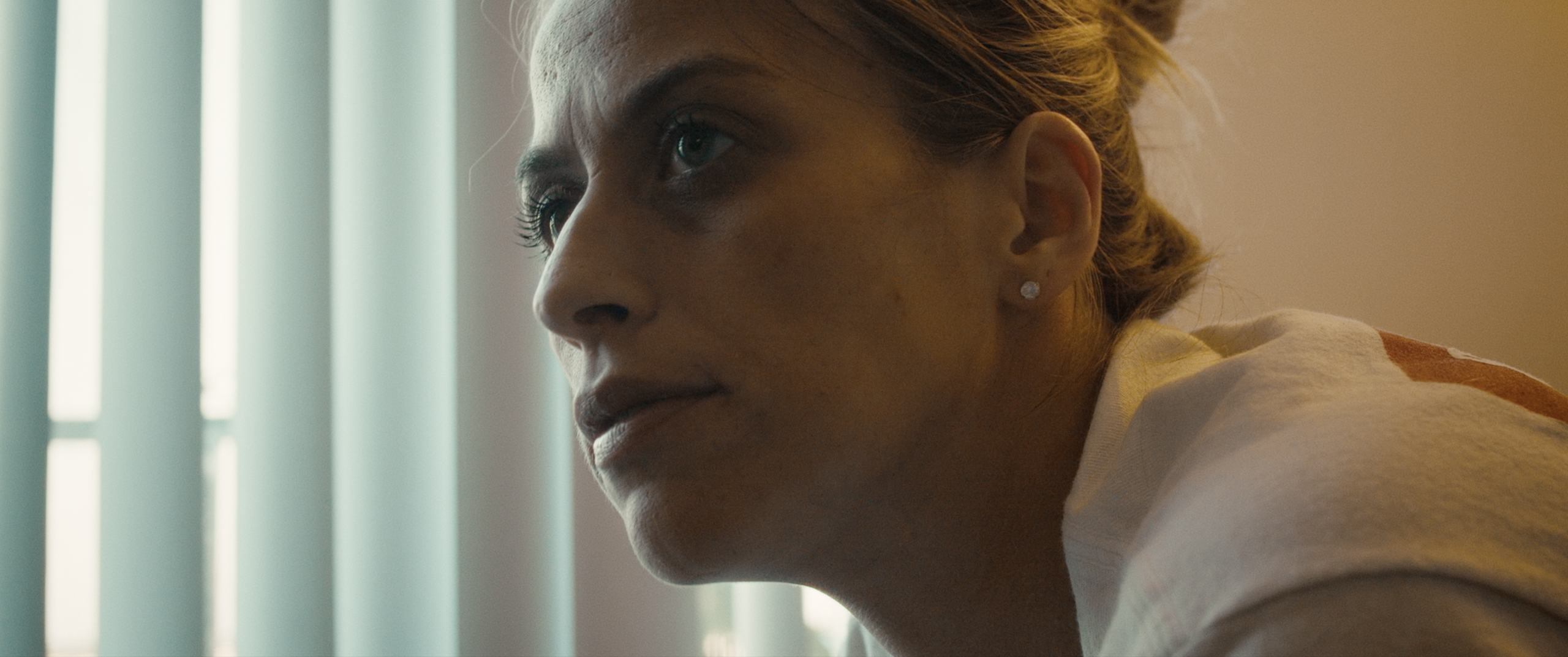 A woman with pulled-back hair and stud earrings looks to the side in a softly lit room with vertical blinds in the background.