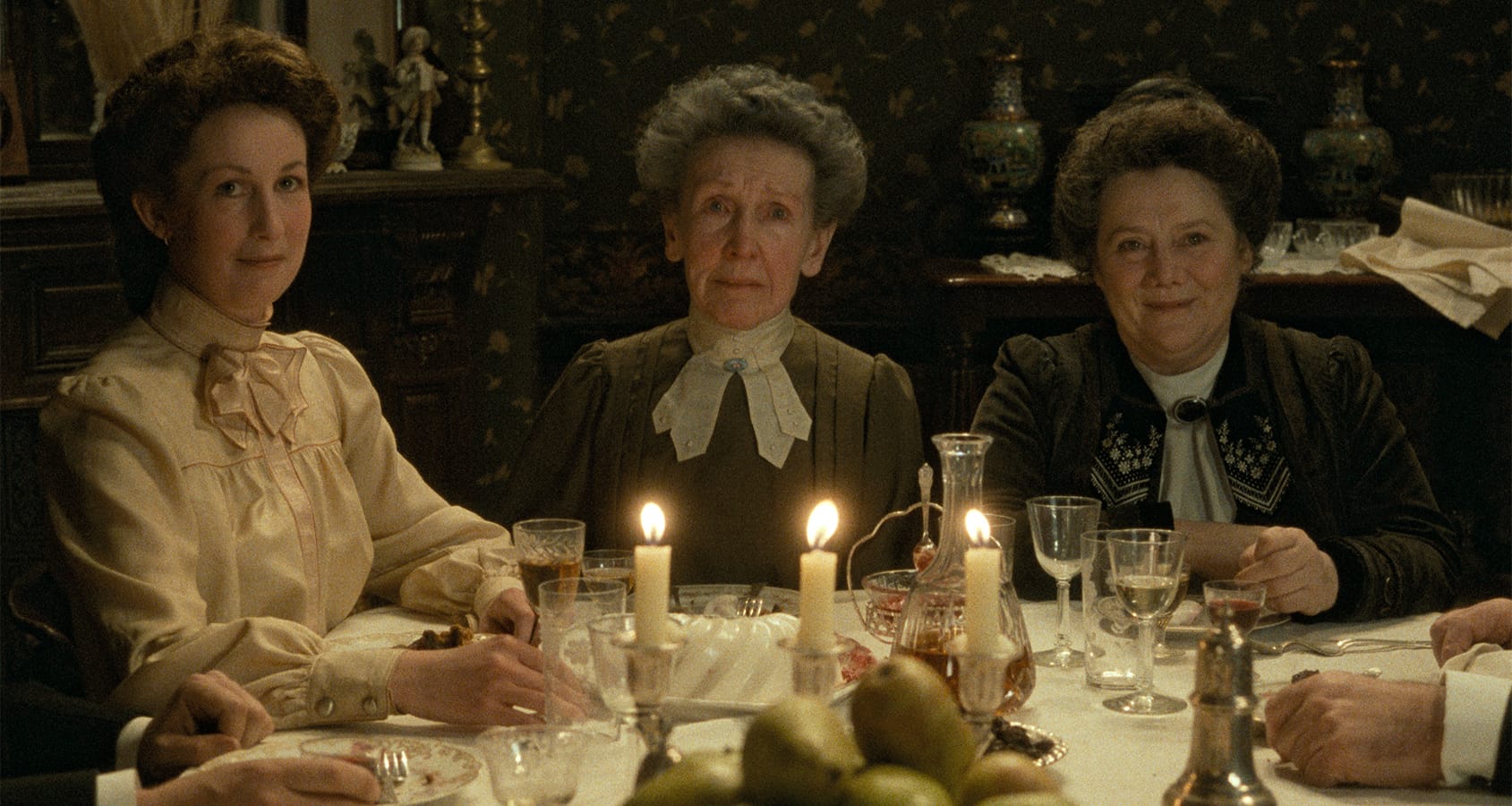 Three older women in period clothing sit at a candlelit dining table set with glasses, fruit, and dishes, facing the camera in a dimly lit, vintage room.