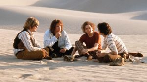 Four people sit on sand in a desert, facing each other in a circle, engaged in conversation, with shoes placed nearby.