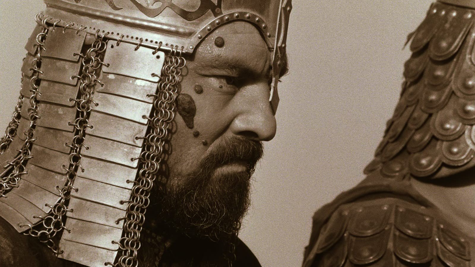 A man wearing an ornate metal helmet and armor, with facial markings and a serious expression, viewed in profile.