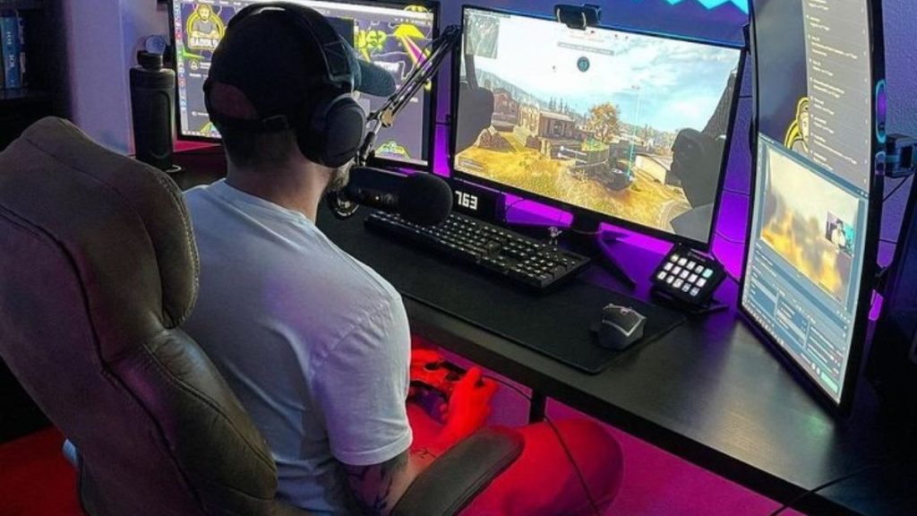 A person wearing headphones sits at a desk with multiple monitors, playing a video game using a controller and streaming equipment.