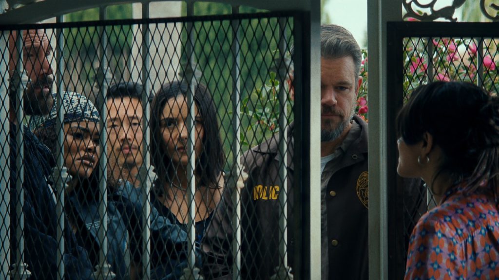 Six people stand behind a metal gate, facing a woman on the other side who appears to be speaking to them. The scene is outdoors with greenery in the background.