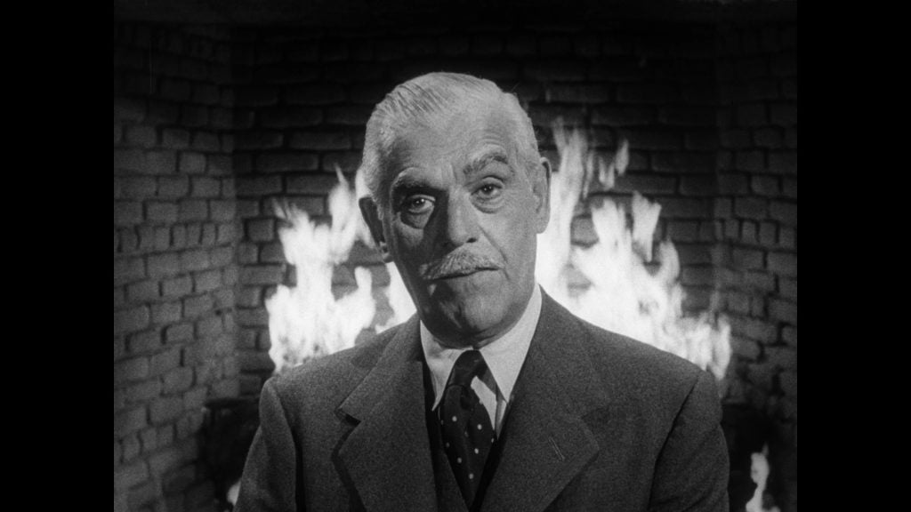 An older man in a suit and tie stands in front of a lit brick fireplace, looking directly at the camera.