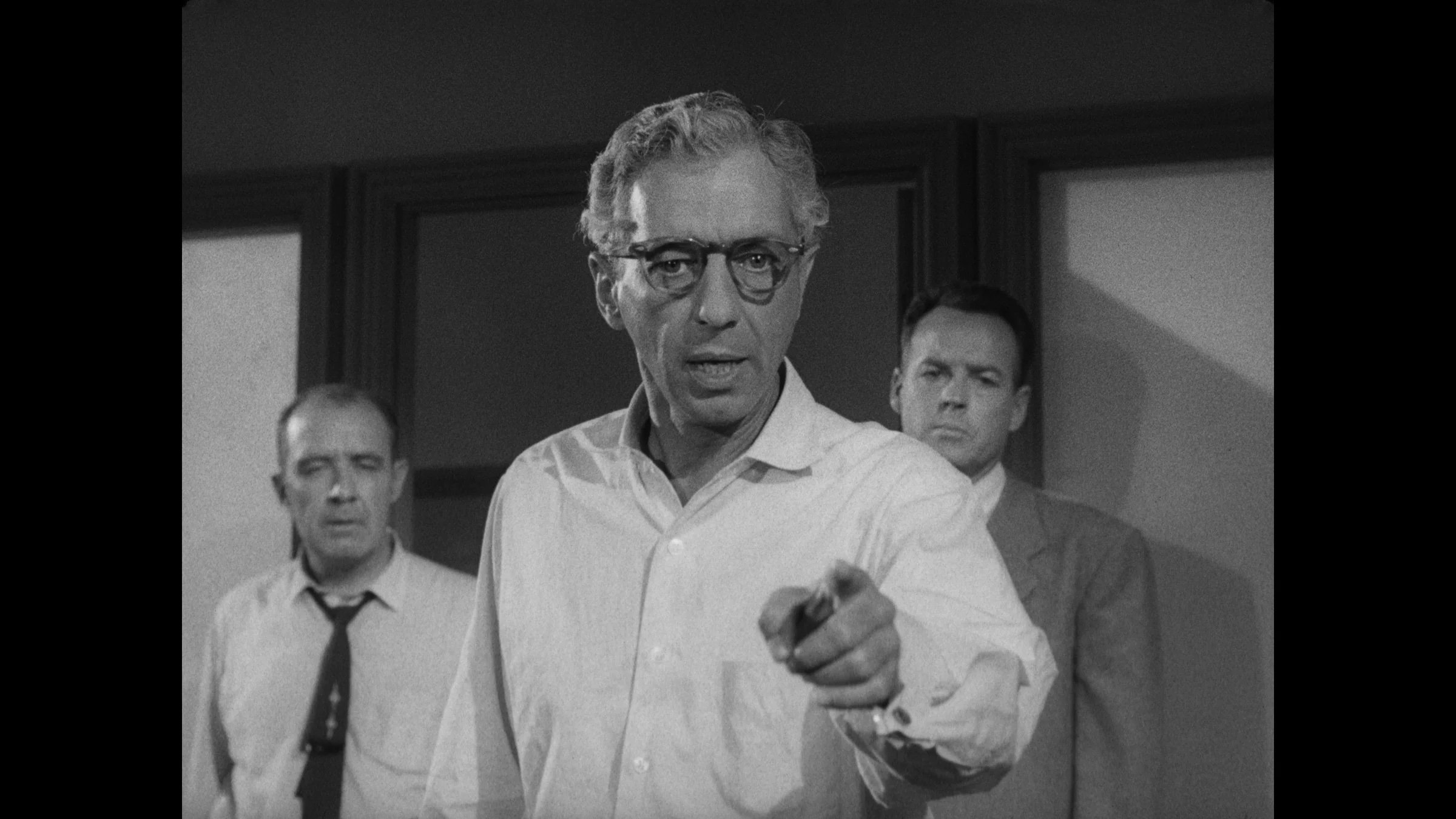 An older man in glasses and a white shirt points forward, with two men standing behind him in a room with dark-paneled walls.