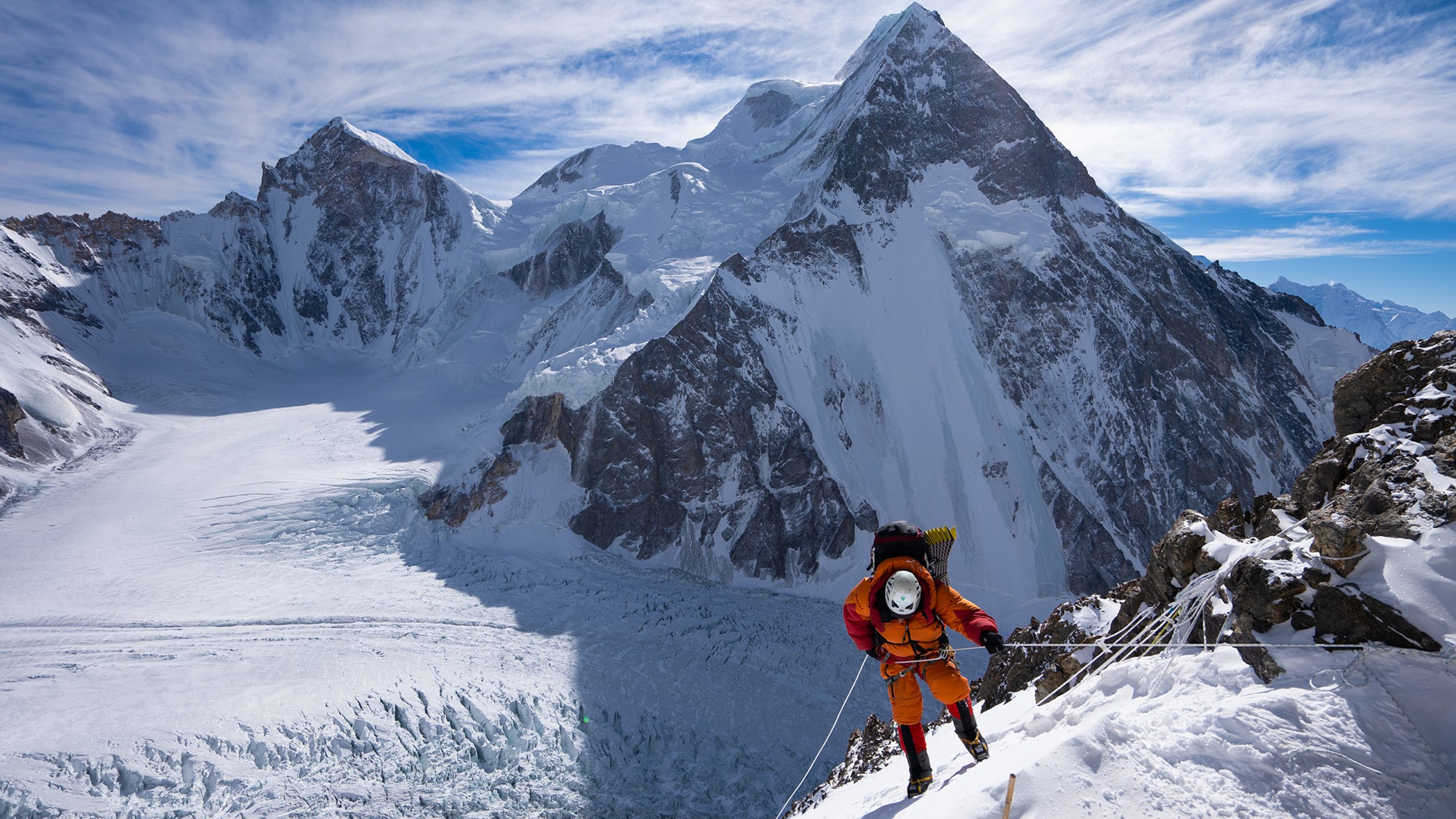 A climber in orange gear ascends a snowy, steep mountain slope using ropes, with large snow-covered peaks and glaciers visible in the background.