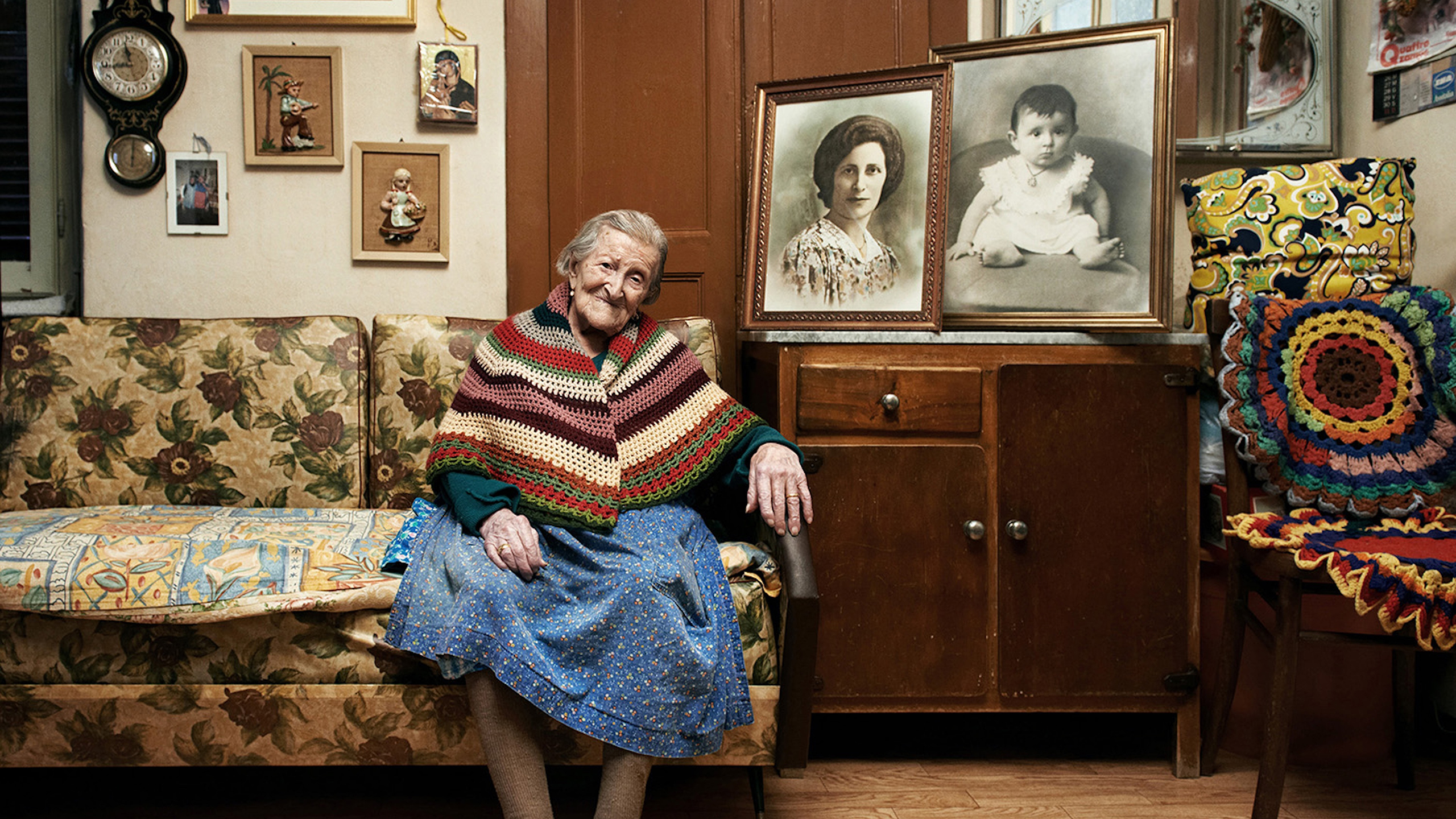 An elderly woman wearing a colorful shawl sits on a floral couch in a cozy room, surrounded by framed family photos and vintage decor.