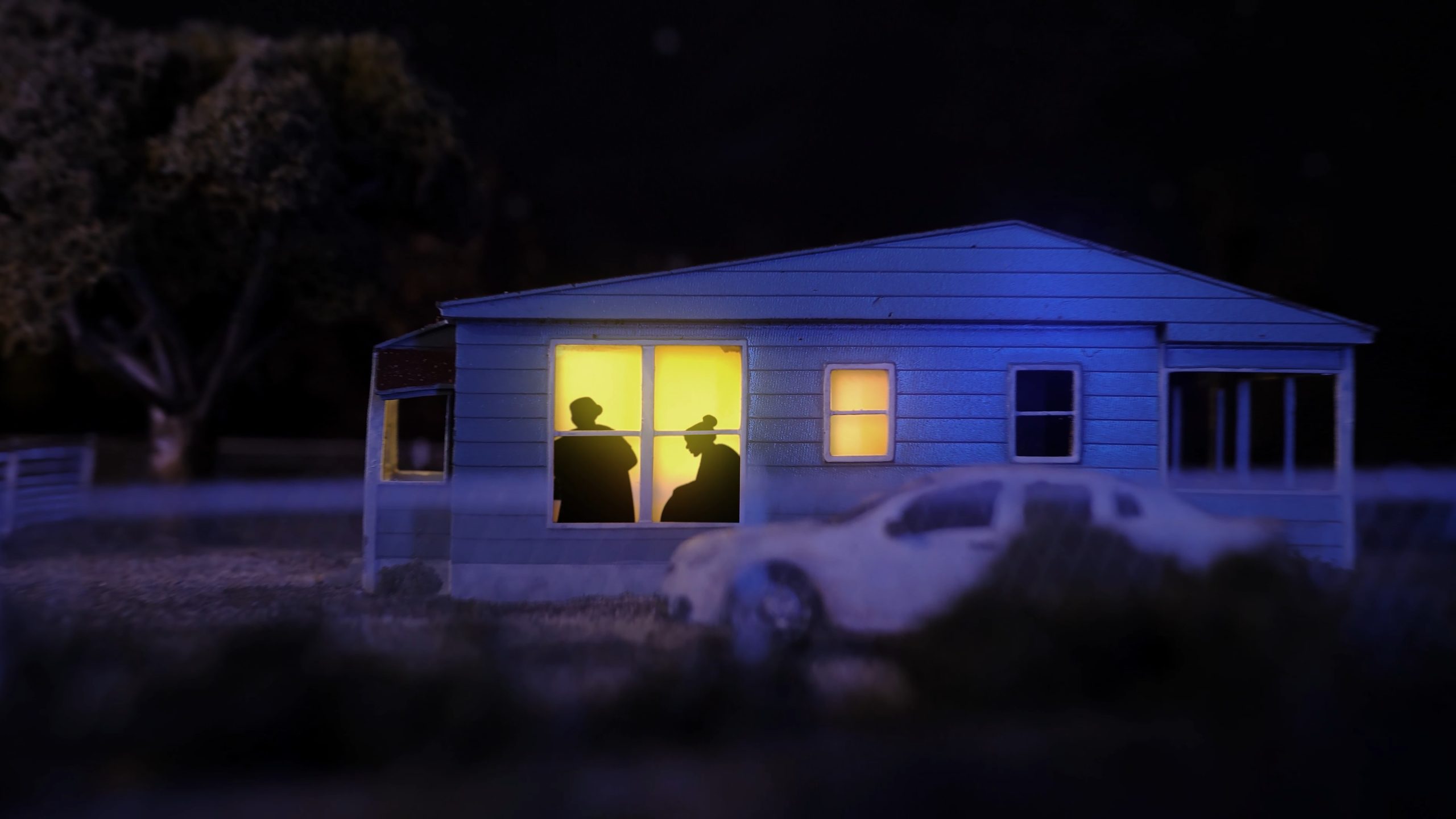 A small blue house at night with two silhouetted figures visible through a brightly lit window; a white car is parked outside.
