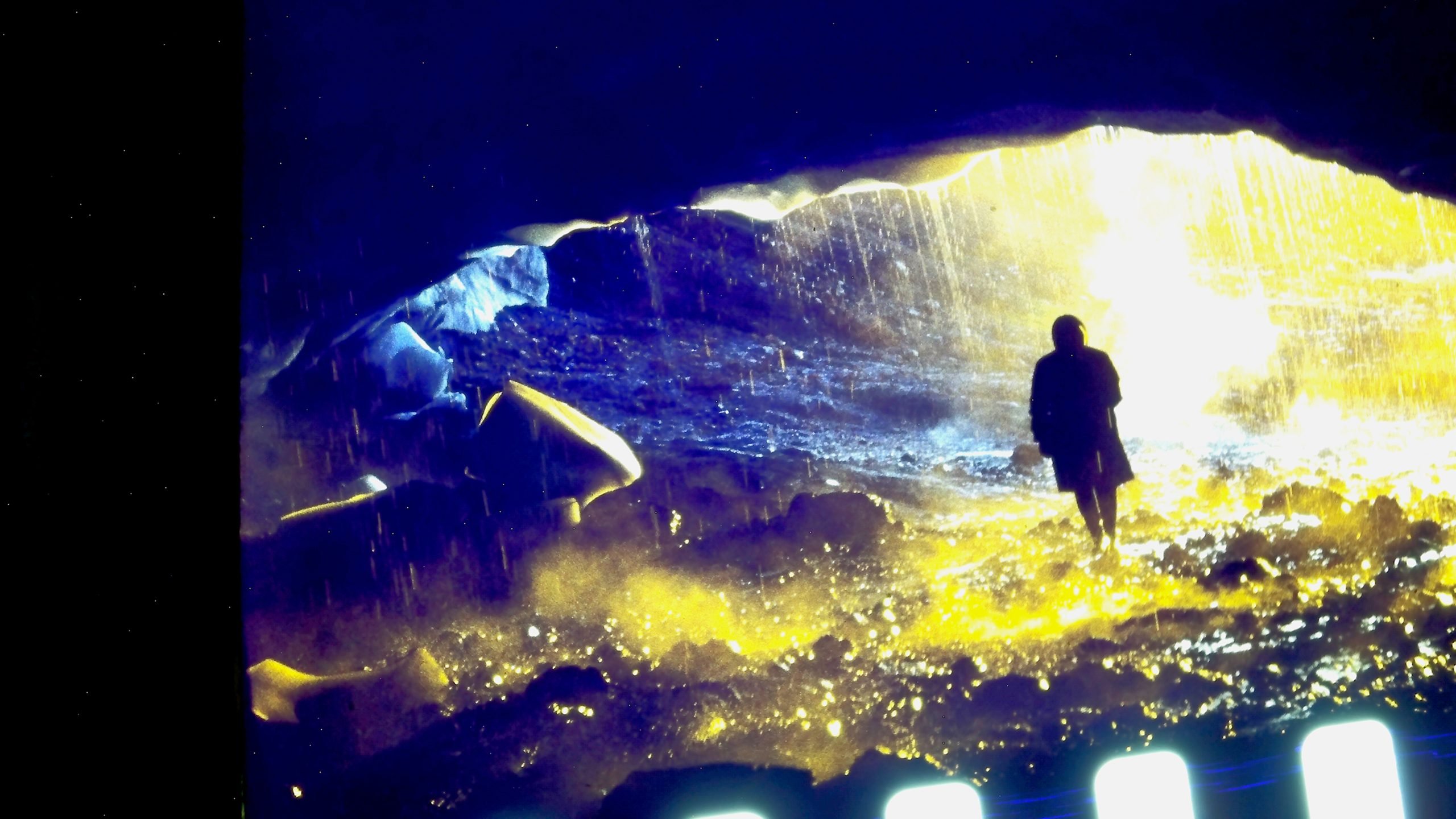 A person stands at the entrance of a cave, illuminated by bright yellow and blue light with water streaming down from above.