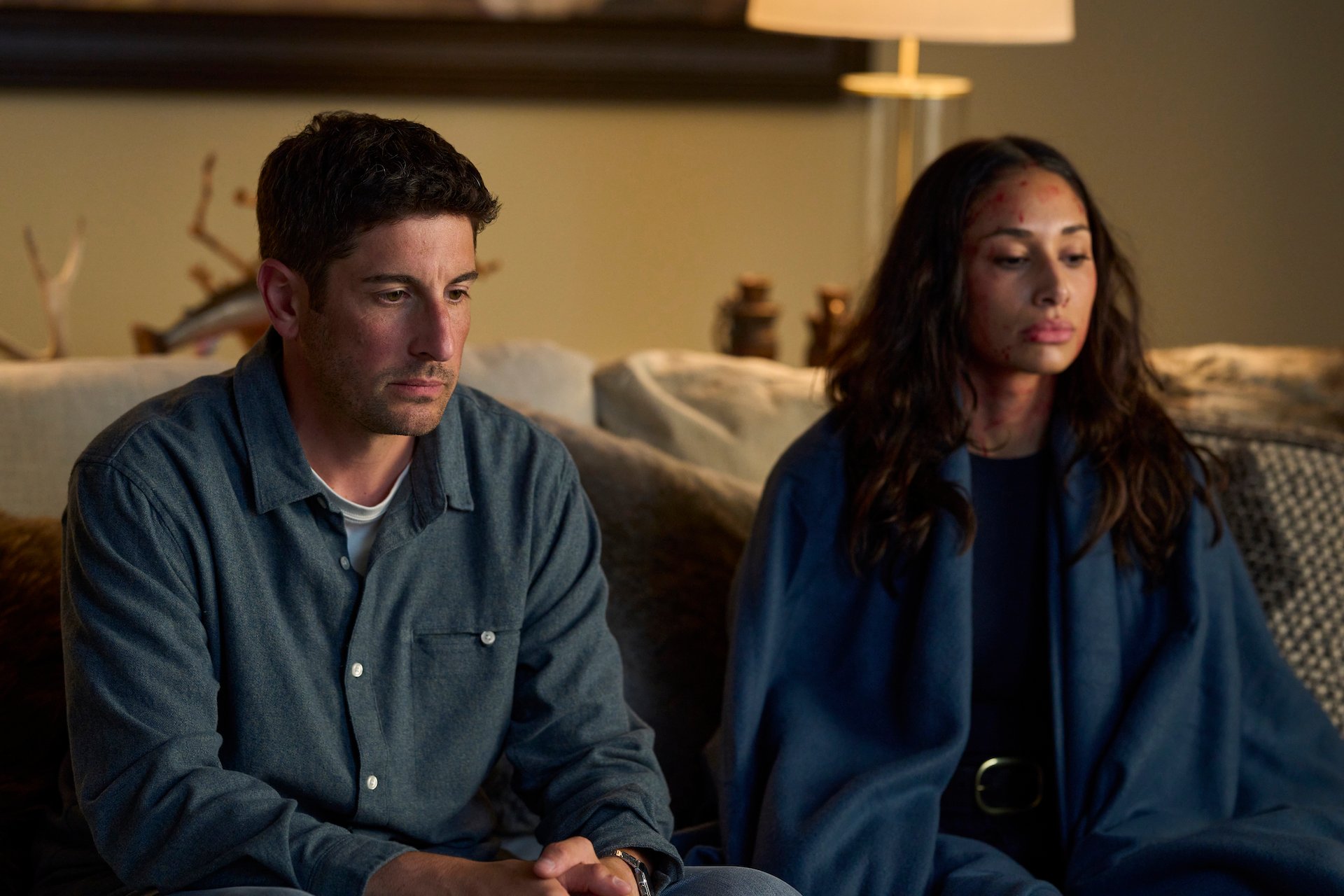 A man and a woman sit side by side on a couch. The woman has visible facial injuries and both appear serious and contemplative.