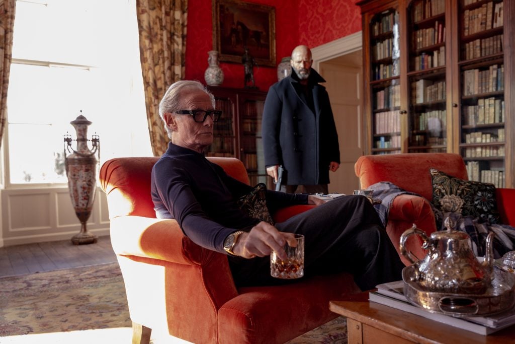 An older man sits in an orange armchair holding a glass of liquor, while another man stands behind him in a room with bookshelves and ornate decor.