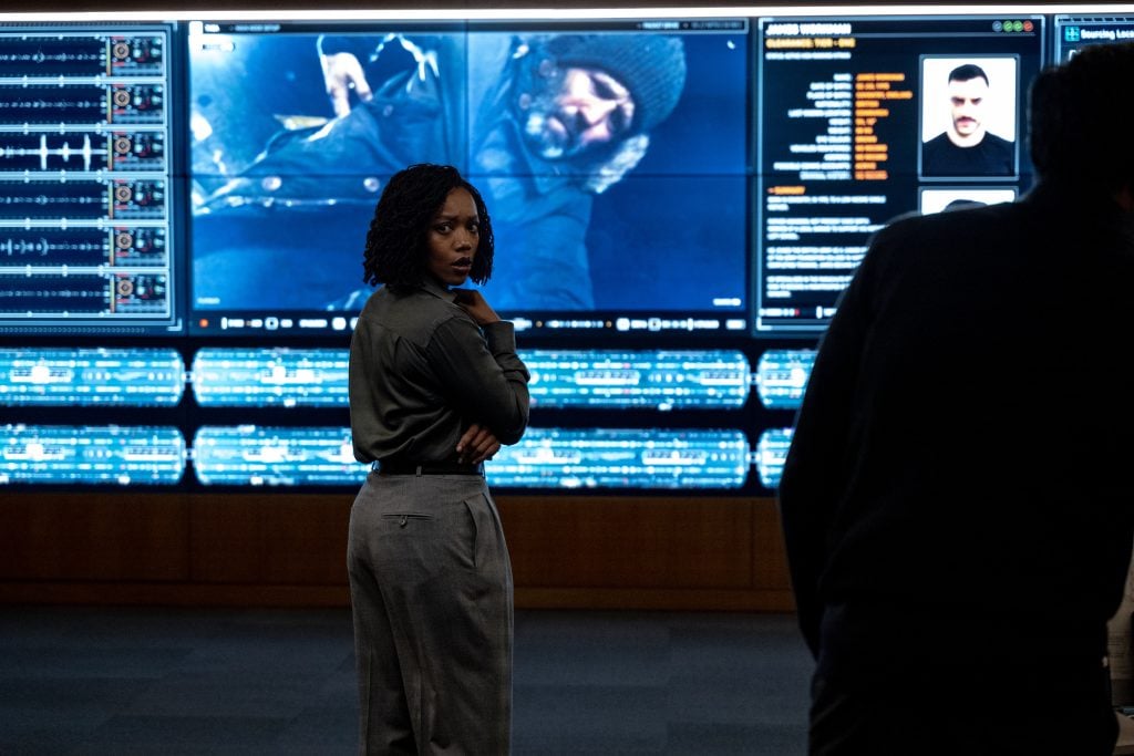 A woman stands in front of large digital screens displaying data, images, and profiles, with another person partially visible in the foreground.