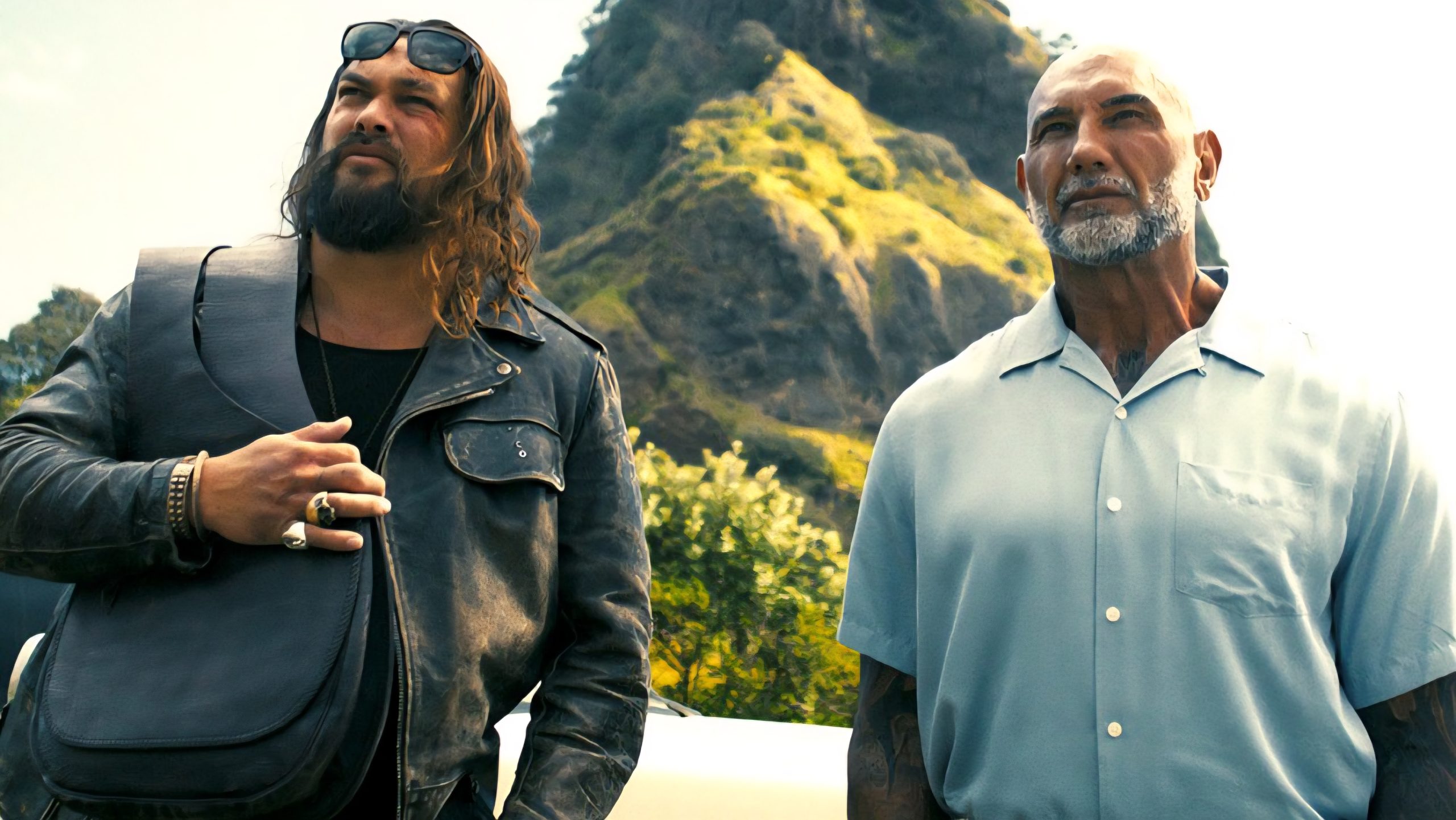 Two men stand outdoors in front of a mountain; one wears a leather jacket and crossbody bag, the other a light short-sleeve shirt.