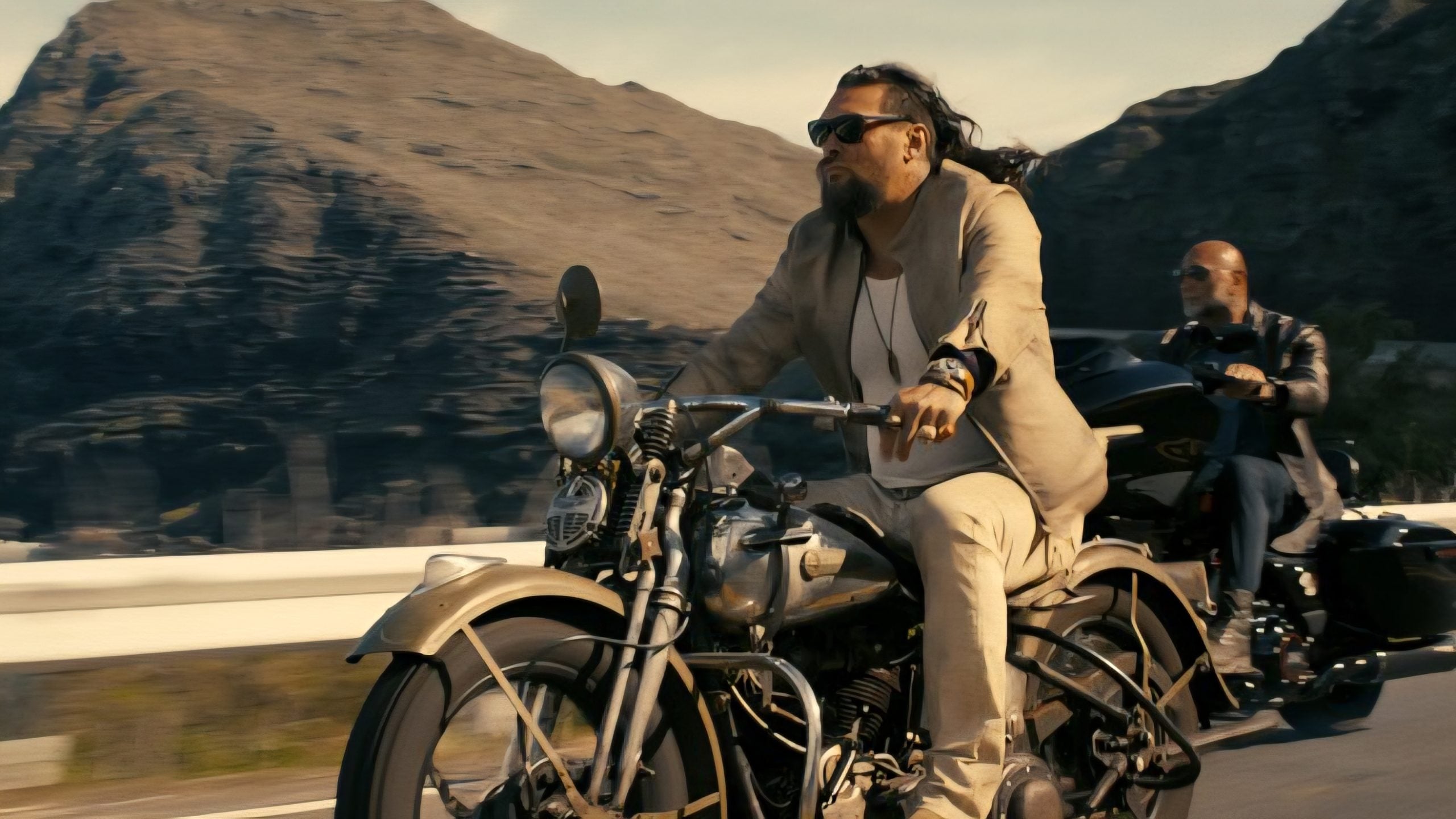 Two men ride motorcycles on a road with rocky mountains in the background; the man in front wears light clothing and sunglasses.