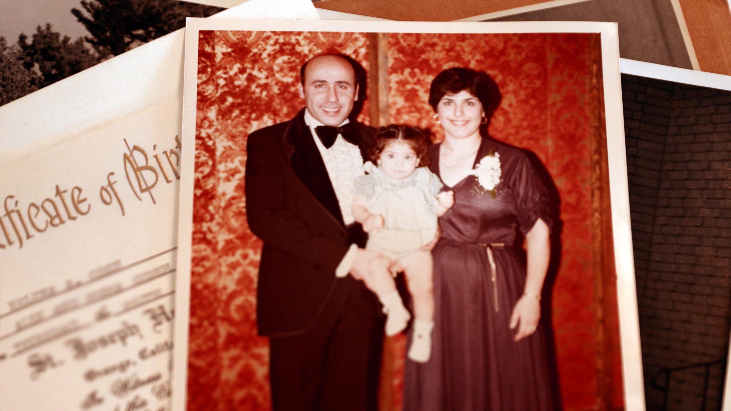 A man in a tuxedo and a woman in a formal dress pose with a young child for a portrait against a patterned red backdrop.