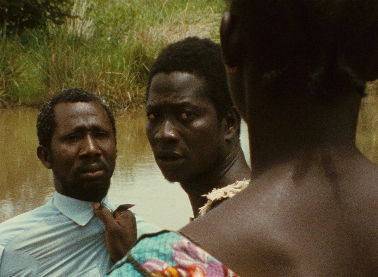 Two men stand by a river, with one holding the other by the collar, while a third person with a colorful garment stands in the foreground facing them.