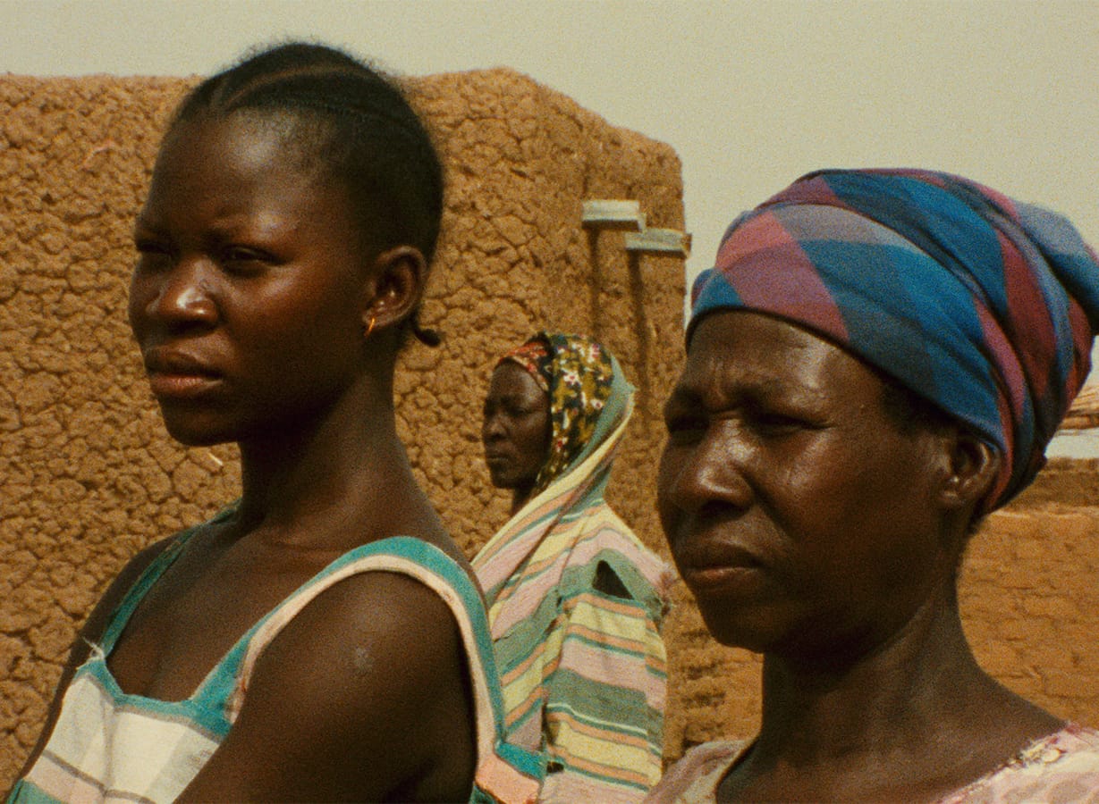 Three women in patterned clothing stand outside near mud-brick buildings, with two in the foreground and one in the background; all appear to be looking in the same direction.