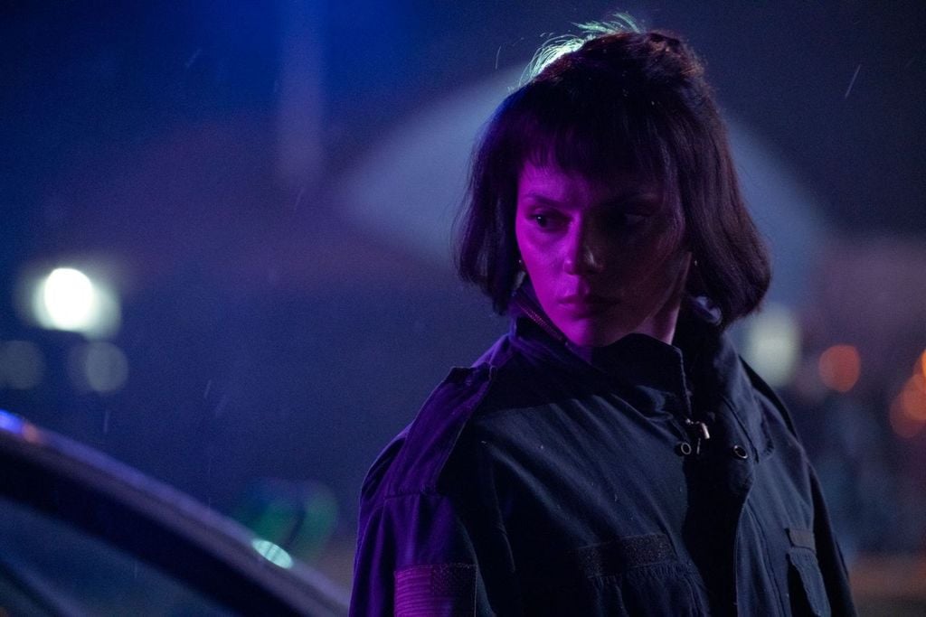 A person with short dark hair and bangs stands outdoors at night under purple and blue light, wearing a dark jacket and looking to the side.