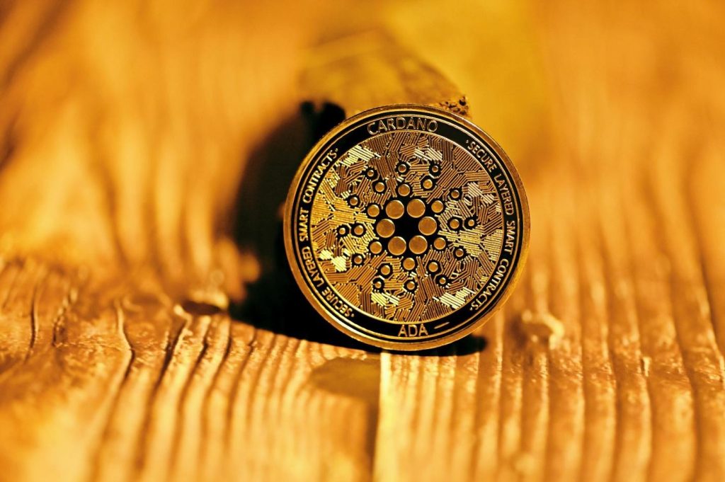 A Cardano ADA cryptocurrency coin rests on a wooden surface, with detailed engravings and circuit-like patterns visible.