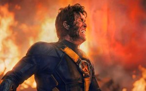 Scott Summers Is Finally Getting The Hype He Deserves A man in a scorched, futuristic suit with an X emblem winces in pain, standing before large flames and smoke in the background.