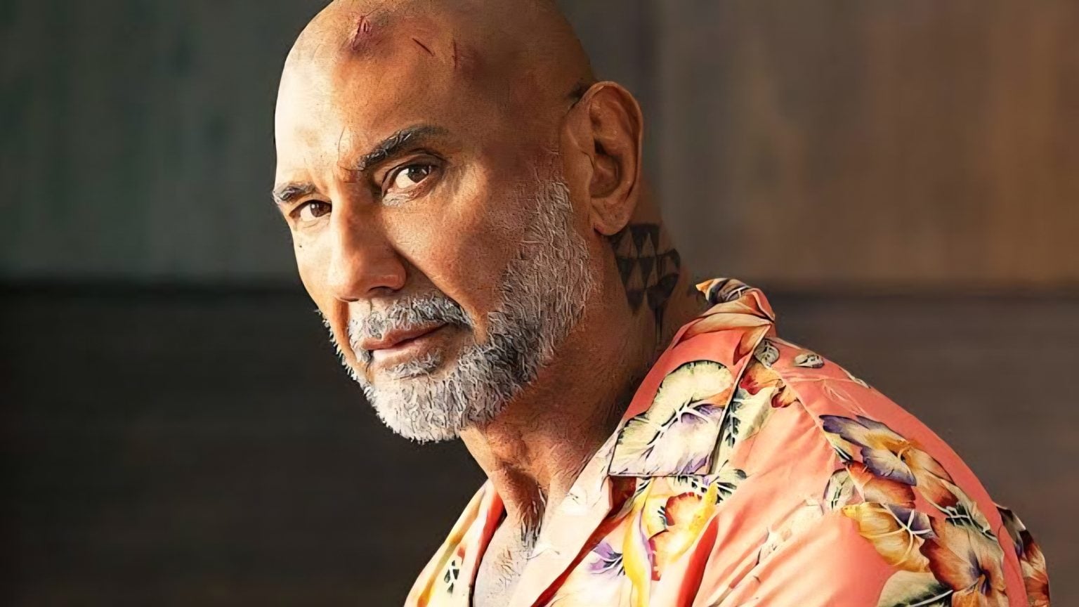 A bald man with a gray beard and visible scars on his head looks toward the camera, wearing a floral shirt with a geometric tattoo on his neck.
