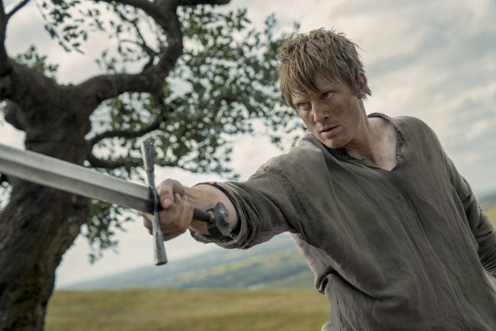 A person with a serious expression holds a sword extended forward, standing outdoors near a tree with hills in the background.