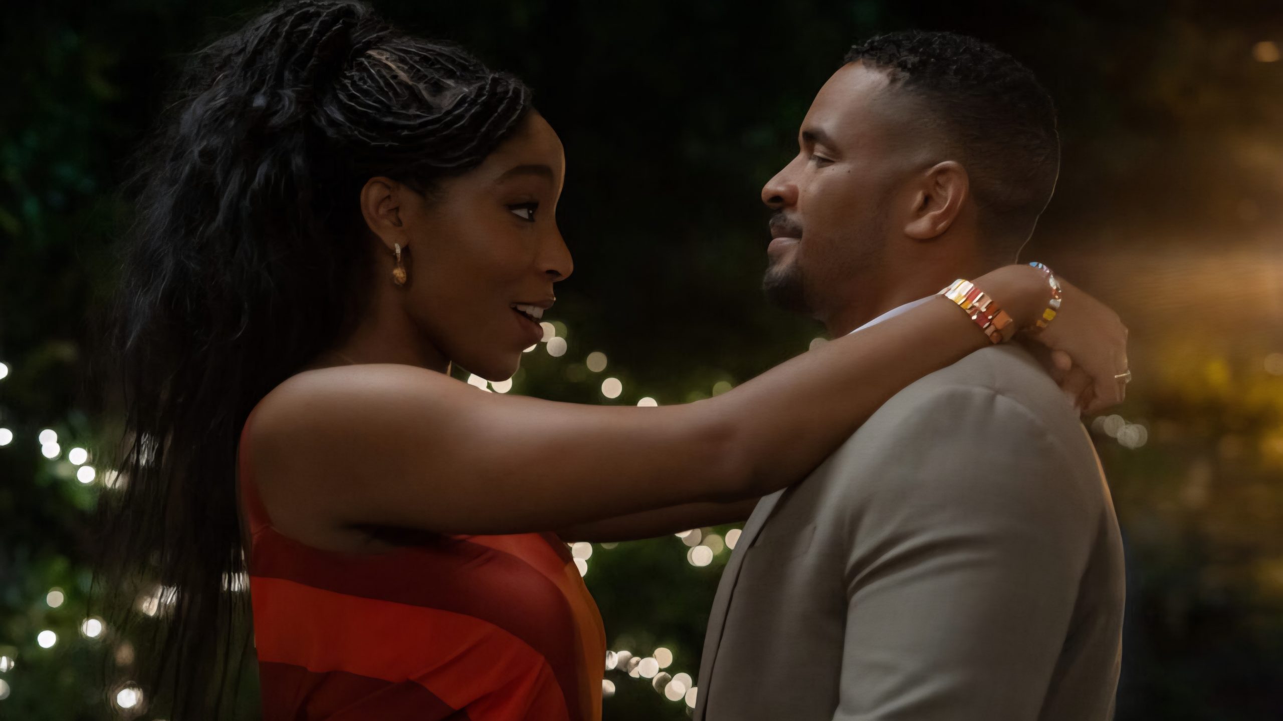 A woman in a red dress and a man in a suit stand close together at night, facing each other with her arms around his neck; string lights are visible in the background.