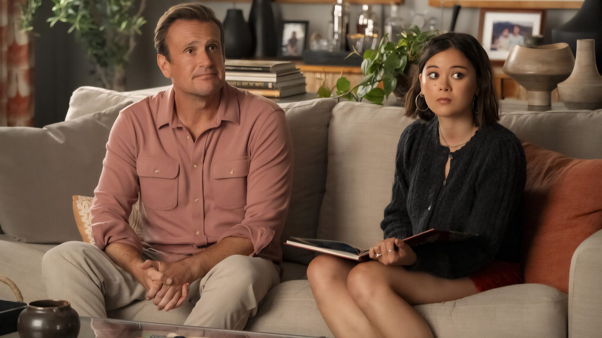 A man and a woman sit on a beige sofa, the man in a pink shirt and the woman holding a book, looking slightly off-camera in a warmly lit living room.