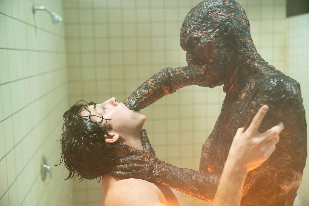 A monstrous, burned-looking creature forces its arm into a man's mouth while holding his neck in a tiled shower room.