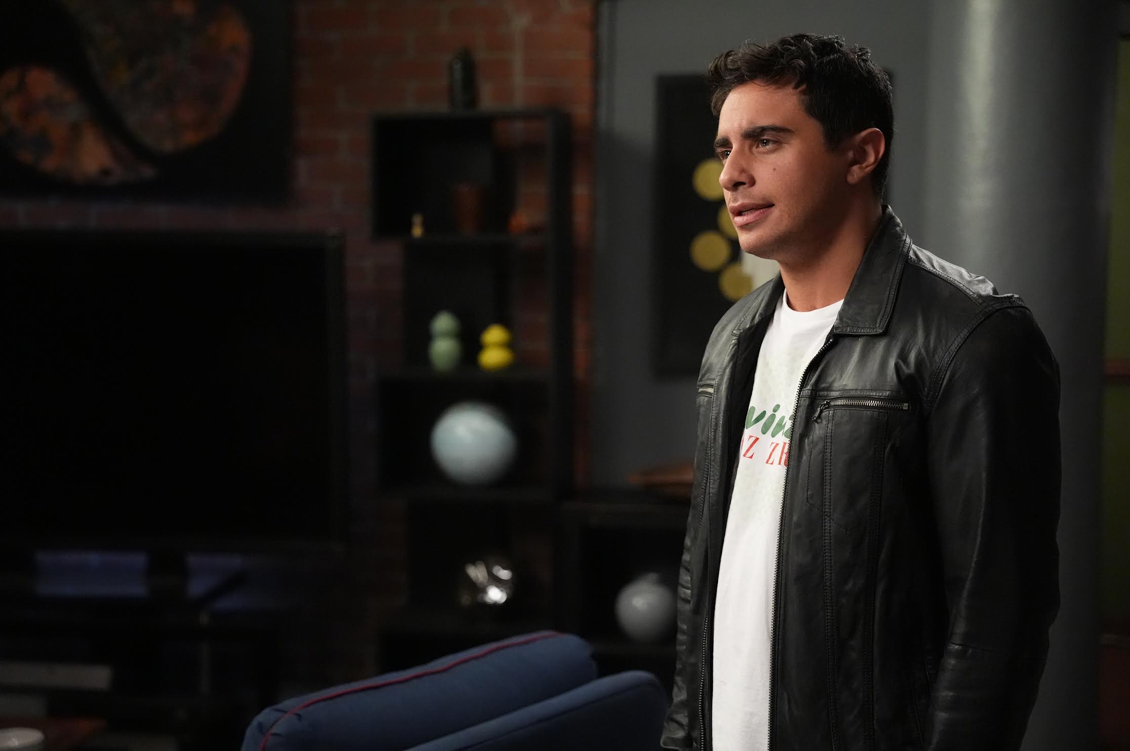 A man in a black leather jacket and white t-shirt stands indoors in a modern living room with shelves, decor, and a TV in the background.